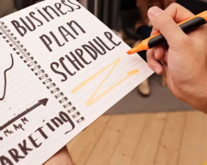 A hand writing the words business plan schedule and marketing in a spiral notebook with an orange marker